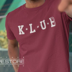 Op de afbeelding: Een maroon t-shirt met witte tekst die "K*L*U*B" zegt.