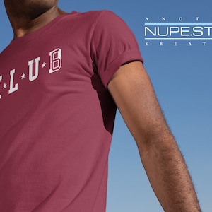 Op de afbeelding: Een maroon t-shirt met witte letters die "K-L-U-B" lezen. De tekst is in een vintage universitaire stijl lettertype. Het t-shirt wordt gedragen door een persoon die voor een blauwe lucht staat.