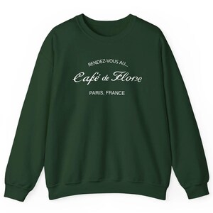 May include: A dark forest green crewneck sweatshirt featuring white text printed on the chest. The text reads RENDEZ-VOUS AU... Café de Flore PARIS, FRANCE in a combination of sans-serif and elegant cursive script lettering.