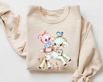 Peut inclure: Un sweat-shirt beige avec une illustration vintage d'un agneau et d'un ours en peluche. L'agneau porte une guirlande de fleurs et l'ours en peluche tient un oiseau bleu.