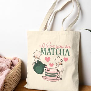 Puede incluir: Bolso tote color crema con el texto "I love you so MATCHA" e ilustraciones de dos figuras de querubines. Una figura vierte de un recipiente verde en una taza de t&eacute; a rayas rosas y verdes. Tambi&eacute;n se presentan corazones rosas.