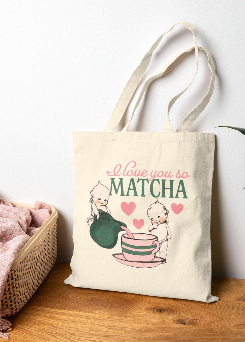 Puede incluir: Bolso tote color crema con el texto "I love you so MATCHA" e ilustraciones de dos figuras de querubines. Una figura vierte de un recipiente verde en una taza de t&eacute; a rayas rosas y verdes. Tambi&eacute;n se presentan corazones rosas.