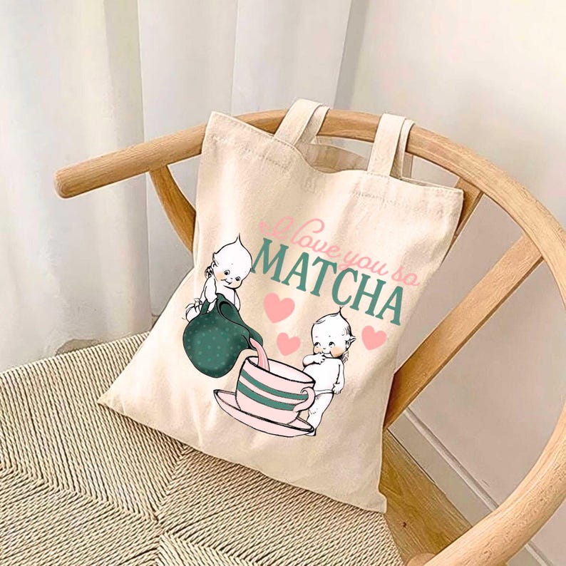 Puede incluir: Bolso tote de lona beige con el texto "I love you so MATCHA" en rosa y verde. El bolso presenta un gr&aacute;fico de dos figuras de querubines, una vertiendo de una jarra verde en una taza de t&eacute; rosa, con corazones rosas.