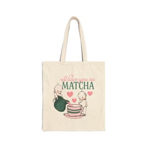 Puede incluir: Bolso tote color crema con la frase "I love you so MATCHA" y un dise&ntilde;o de dos figuras de querubines, una tetera, una taza de t&eacute; y corazones rosas. El bolso es rectangular y tiene dos asas. El dise&ntilde;o es verde y rosa.