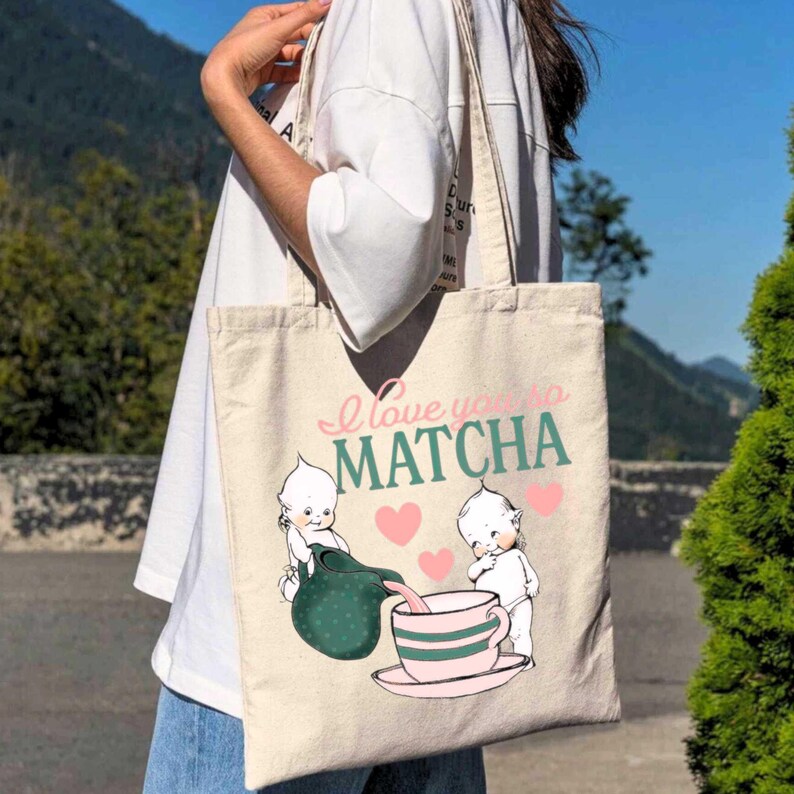 Puede incluir: Bolso tote beige con el texto "I love you so Matcha" en rosa y verde. El bolso presenta un gr&aacute;fico de dos figuras querubinescas vertiendo matcha en una taza de t&eacute; rosa, con corazones rosas.