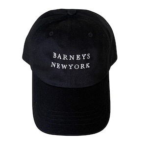 May include: Black baseball cap with the text "BARNEYS NEWYORK" embroidered in white. The cap has a curved brim and a button on top. A classic, casual accessory.