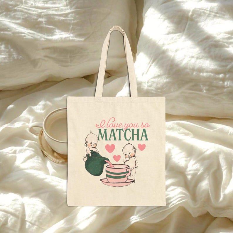 Puede incluir: Bolso tote de color crema con una ilustraci&oacute;n caprichosa. El dise&ntilde;o presenta dos figuras querubinas vertiendo matcha en una taza de t&eacute;, con el texto "I love you so MATCHA" en rosa y verde. El bolso tiene una forma simple y rectangular con dos asas.