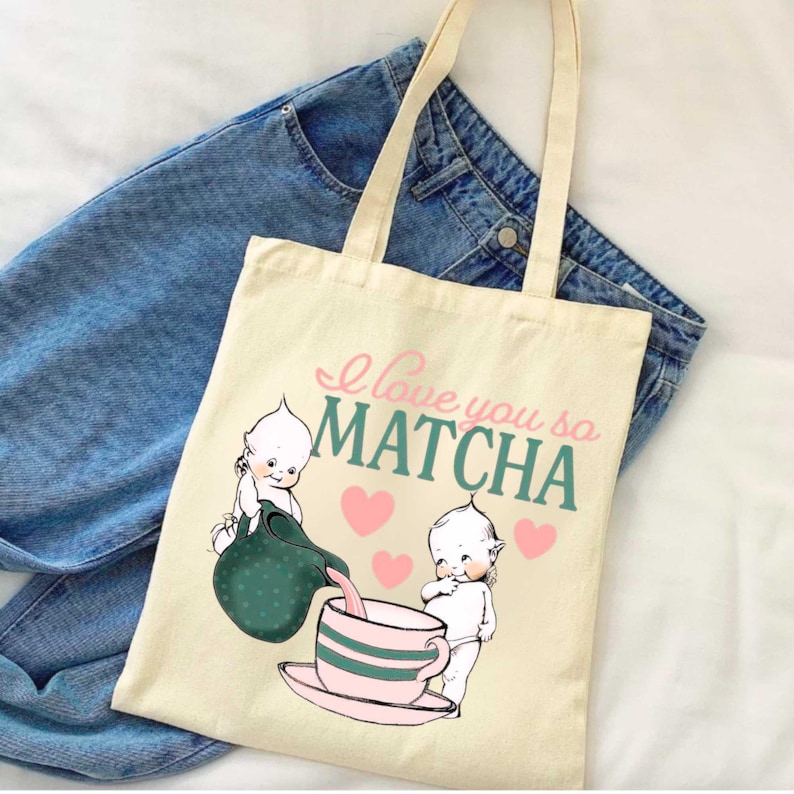 Puede incluir: Bolso tote color crema con el texto "I love you so MATCHA" en rosa y verde azulado. El bolso presenta una ilustraci&oacute;n de dibujos animados de dos figuras de querubines, una tetera y una taza de t&eacute; con corazones rosas.