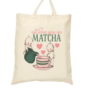 Puede incluir: Bolso tote de color crema con el texto "I love you so MATCHA" en rosa y verde. El bolso presenta una ilustraci&oacute;n vintage de dos figuras querubinescas vertiendo matcha en una taza de t&eacute; rosa. Corazones rosas est&aacute;n dispersos alrededor de las figuras.