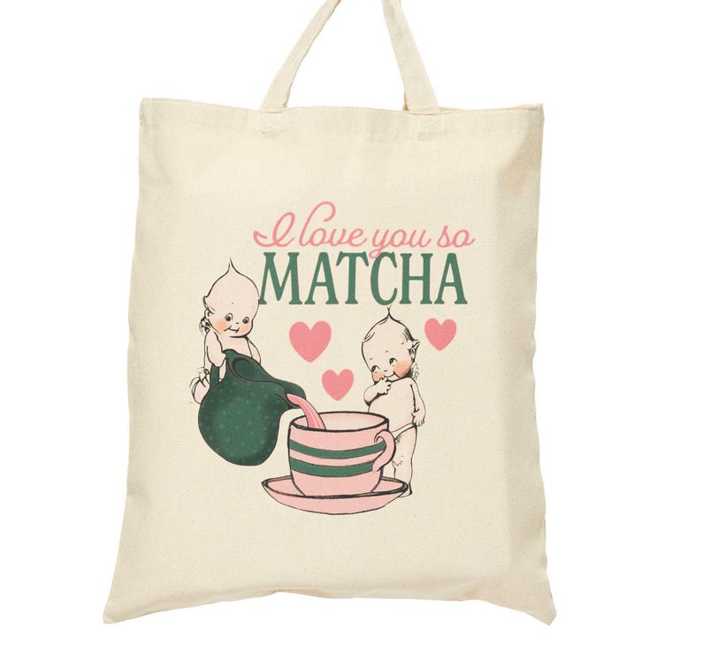 Puede incluir: Bolso tote de color crema con el texto "I love you so MATCHA" en rosa y verde. El bolso presenta una ilustraci&oacute;n vintage de dos figuras querubinescas vertiendo matcha en una taza de t&eacute; rosa. Corazones rosas est&aacute;n dispersos alrededor de las figuras.