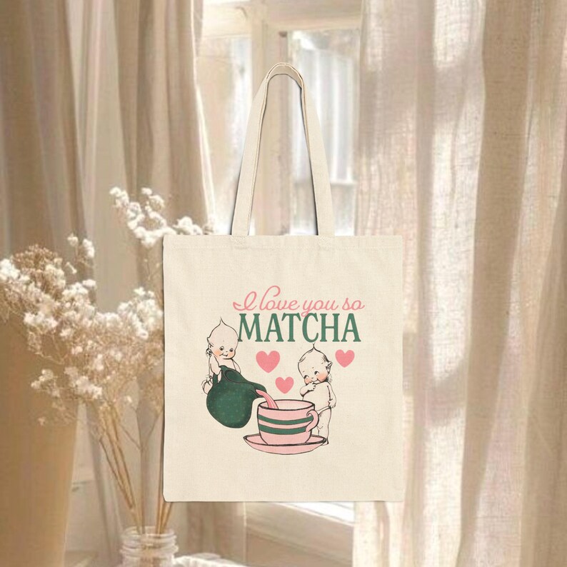 Puede incluir: Bolso tote color crema con el texto "I love you so MATCHA" en rosa y verde. El bolso presenta una ilustraci&oacute;n de dos figuras querubinescas vertiendo matcha en una taza de t&eacute;, con corazones rosas.