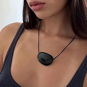 Puede incluir: Un colgante de piedra negra pulida y lisa suspendido de un fino cordón negro. El colgante tiene una forma irregular y redondeada con una superficie brillante y reflectante que capta la luz, creando una estética sencilla y limpia para la joya.