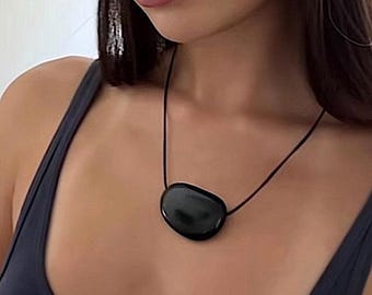 May include: A smooth, polished black stone pendant suspended on a thin black cord necklace. The pendant has an irregular, rounded shape with a glossy, reflective surface that catches light, creating a simple and clean aesthetic for the jewelry piece.
