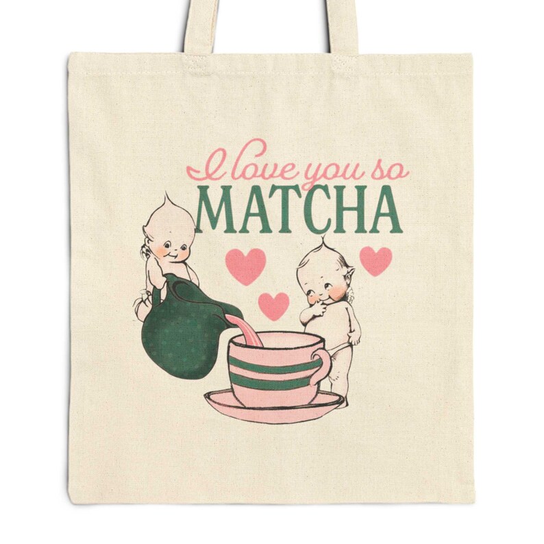 Puede incluir: Bolso tote color crema con una ilustraci&oacute;n de estilo vintage. El dise&ntilde;o presenta dos figuras de querubines, una tetera vertiendo en una taza a rayas y el texto "I love you so MATCHA" en rosa y verde. Corazones rosas dispersos.