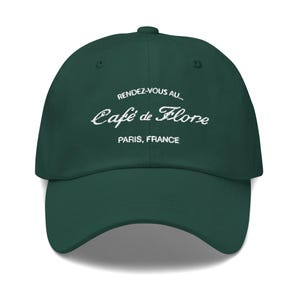 Puede incluir: Una gorra de béisbol de color verde oscuro con texto bordado en blanco que dice RENDEZ-VOUS AU... Café de Flore PARIS, FRANCE. La gorra tiene una corona estructurada, una visera curva y un botón en la parte superior, diseño de seis paneles.