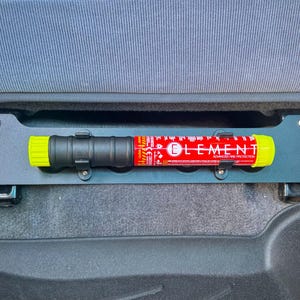 May include: A black metal bracket with a fire extinguisher, mounted under a car seat. The fire extinguisher is red with yellow ends and the word "ELEMENT" printed on it. The bracket is secured with bolts.