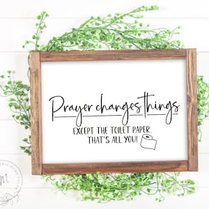 以下が含まれることがあります： 茶色の木製の枠が付いた白い看板で、"祈りは物事を変える。トイレットペーパー以外はね。それだけだよ！"というテキストと、漫画のトイレットペーパーのロールのイラストが描かれている。