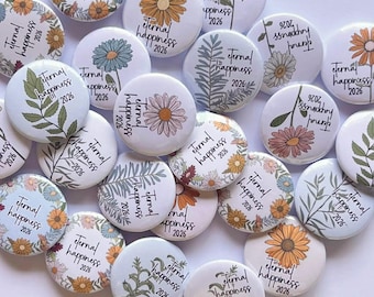 May include: A collection of white, round buttons with floral designs and the text "eternal happiness 2026." The buttons feature various flower illustrations in shades of yellow, orange, blue, and pink, along with green foliage.