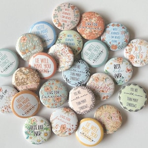 Op de afbeelding: Een assortiment van kleurrijke ronde knoppen met inspirerende citaten en bloemmotieven. De citaten omvatten "Geloof kan dingen laten gebeuren", "Paradijs", "Hij zal je sterk maken", "Voor altijd leven", "Je bent meer waard dan veel vogels", en "Het beste leven ooit".