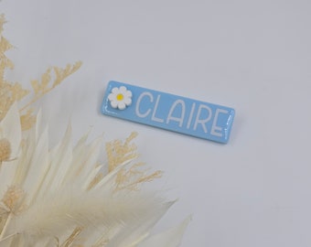 May include: A light blue rectangular name tag with a white daisy and the name "CLAIRE" in white letters.