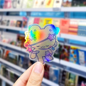 May include: Holographic sticker of a cartoon bear with a rainbow head and stars. The bear has a dreamy expression and is surrounded by star-shaped accents. The sticker is held in front of a blurred background of bookshelves.