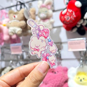 May include: A clear acrylic keyring with a cartoon bear design, pink bow tie, hearts, stars, and smaller bear figures. The keyring is attached to a silver clasp, with pink and blue accents.