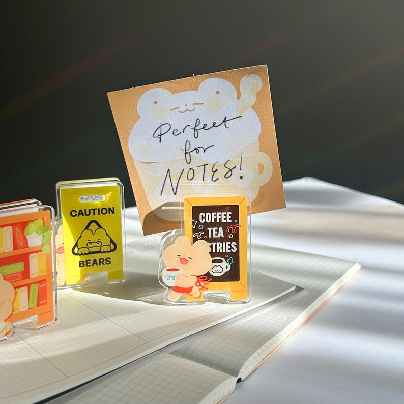 Puede incluir: Colecci&oacute;n de accesorios de escritorio acr&iacute;licos con dise&ntilde;os de osos de dibujos animados. Incluye un soporte para blocs de notas con el texto "Perfect for NOTES!", una se&ntilde;al de precauci&oacute;n y un letrero de cafeter&iacute;a. Sobre un cuaderno cuadriculado.