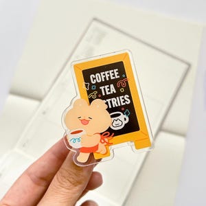 Puede incluir: Una pegatina acr&iacute;lica transparente con un oso de dibujos animados sosteniendo una taza de caf&eacute;. El oso est&aacute; frente a un letrero amarillo que dice "Coffee Tea & Tries". La pegatina tiene un dise&ntilde;o de cinta roja.