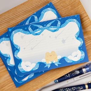 Könnte beinhalten: Blaue Schreibwaren mit einem skurrilen Design. Die Schreibwaren haben einen hellblauen Rand mit einer Schleife, Sternschnuppen und einem gelben Frosch auf einer Wolke. Die Schreibwaren sind liniert und haben einen weißen Bereich zum Schreiben. Der Text "@soogstudio" befindet sich unten rechts.