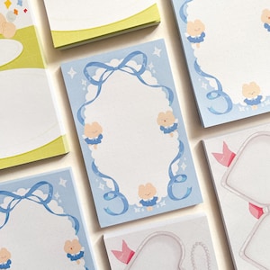 Könnte beinhalten: Ein Set aus blauen und weißen Notizblöcken mit einem Wolken- und Schleifendesign. Jeder Block hat einen weißen Rand mit einem gepunkteten Muster und Illustrationen von kleinen Tieren, die blaue Kleider tragen. Der Text "Happy" ist auf einem der Blöcke gedruckt.