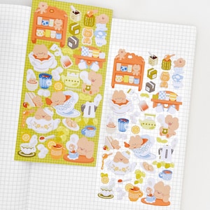 May include: Two sticker sheets with a tea party theme. The stickers feature cartoon bears, teacups, teapots, and small shelves. The color palette includes orange, blue, white, and yellow. The stickers are on a green background.