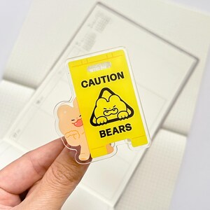 Puede incluir: Una pegatina acr&iacute;lica transparente con un letrero de precauci&oacute;n amarillo con las palabras "CAUTION BEARS" y una ilustraci&oacute;n de oso de dibujos animados. Un personaje de oso marr&oacute;n claro se asoma por detr&aacute;s del letrero.