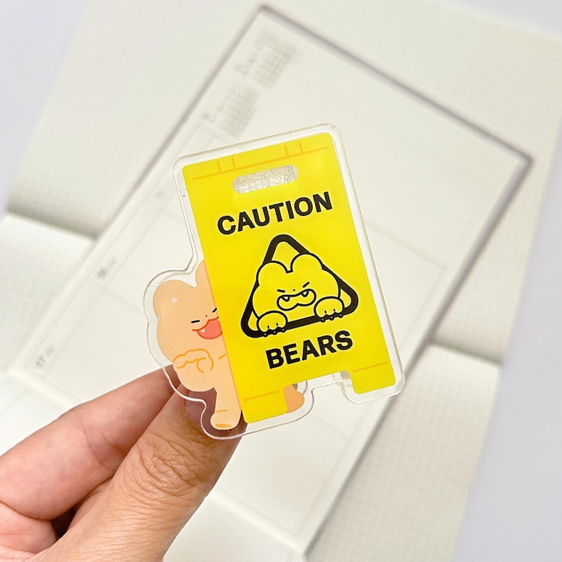 Puede incluir: Una pegatina acr&iacute;lica transparente con un letrero de precauci&oacute;n amarillo con las palabras "CAUTION BEARS" y una ilustraci&oacute;n de oso de dibujos animados. Un personaje de oso marr&oacute;n claro se asoma por detr&aacute;s del letrero.
