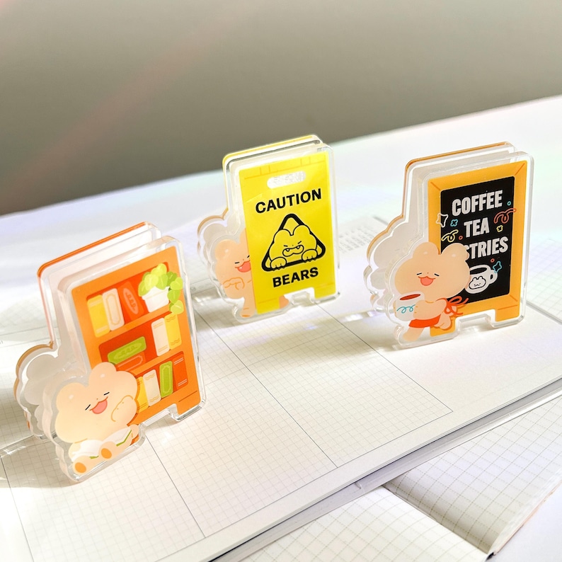 Puede incluir: Tres accesorios de escritorio acr&iacute;licos con dise&ntilde;os de osos de dibujos animados. Uno es una estanter&iacute;a, otro un letrero de precauci&oacute;n con un oso y el tercero un letrero de cafeter&iacute;a. Los accesorios son de color naranja, amarillo, negro y blanco.