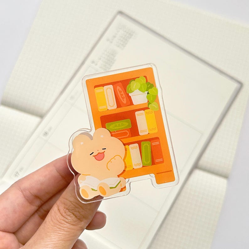 Puede incluir: Una pegatina acr&iacute;lica transparente con un personaje de oso sonriente junto a una estanter&iacute;a naranja. La estanter&iacute;a est&aacute; llena de libros coloridos y una planta en maceta. La pegatina est&aacute; sujeta por una mano.