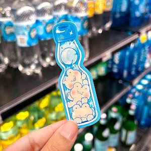 May include: A blue sticker in the shape of a Ramune soda bottle, featuring three cartoon frogs inside. The sticker has a light blue border and a bubbly design, with a star on the top.
