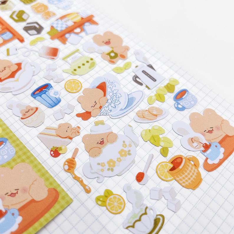 May include: A sheet of stickers featuring a tea party theme. The stickers include images of teddy bears, teacups, teapots, cookies, lemons, and honey. The color palette is soft, with pastel shades of orange, blue, yellow, and white.