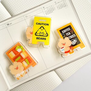 Puede incluir: Tres colgantes acr&iacute;licos transparentes con dise&ntilde;os de osos de dibujos animados. Uno es un letrero de precauci&oacute;n amarillo que dice "CAUTION BEARS". Otro es un letrero de cafeter&iacute;a con "COFFEE TEA TRIES". El &uacute;ltimo es una estanter&iacute;a. Todos est&aacute;n en una agenda.