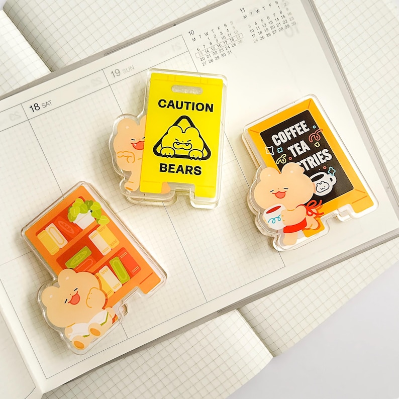 Puede incluir: Tres colgantes acr&iacute;licos transparentes con dise&ntilde;os de osos de dibujos animados. Uno es un letrero de precauci&oacute;n amarillo que dice "CAUTION BEARS". Otro es un letrero de cafeter&iacute;a con "COFFEE TEA TRIES". El &uacute;ltimo es una estanter&iacute;a. Todos est&aacute;n en una agenda.