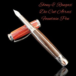 Puede incluir: Una pluma estilográfica con plumín y detalles plateados, y un cuerpo de madera. El capuchón y el cuerpo de la pluma son una mezcla de madera marrón rojiza y marrón oscuro. El texto "Ebony & Rangax Die Cut Atrax Fountain Pen" está en la esquina superior derecha.