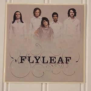 May include: A promotional sticker featuring a band called "FLYLEAF". The sticker has a light pink background with a white border. The band members are wearing white shirts and are standing in front of a light pink background. The band's name is written in a dark red font with a decorative swirl design.