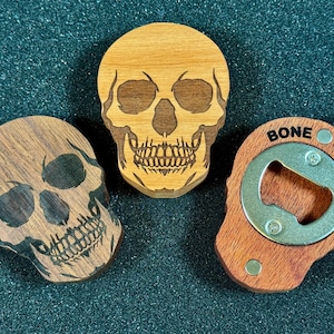 May include: Three wooden skull-shaped items. Two are decorative, with etched skull designs in light and dark wood. The third is a bottle opener, with the words "BONE HEAD" and a metal opener.