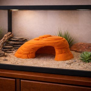 May include: Orange reptile hideout shaped like a rock formation with an arched opening. The hideout is placed inside a terrarium with sand substrate, plants, and other decorative rocks. The terrarium is set on a wooden cabinet.