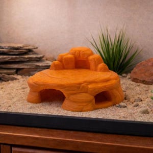 May include: Orange reptile habitat hideout with a rock-like design. The hideout features multiple levels and openings, providing a secure and stimulating environment for reptiles. The hideout is placed on a sandy substrate.