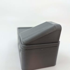 May include: A dark gray rectangular box with a hinged, ridged lid. The box appears to be made of a solid material, possibly plastic or resin. The lid is angled, and the box has a simple, utilitarian design.