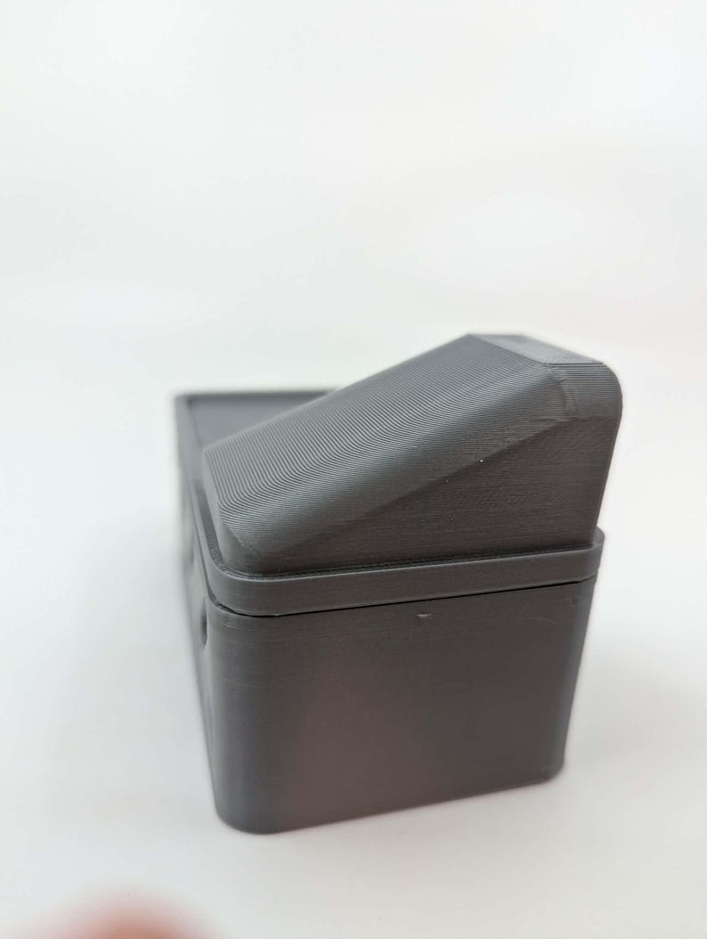 May include: A dark gray rectangular box with a hinged, ridged lid. The box appears to be made of a solid material, possibly plastic or resin. The lid is angled, and the box has a simple, utilitarian design.