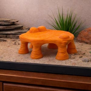 May include: An orange, textured, decorative reptile habitat hideaway. The hideaway is shaped like a small table with four pillar-like legs and a flat top with two rounded protrusions. The hideaway is placed in a terrarium with sand and a few plants.