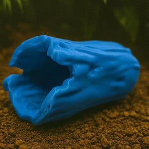 Può includere: Decorazione per acquario a forma di grotta, di colore blu brillante. La struttura cava e strutturata è progettata per fornire riparo alla vita acquatica. L'oggetto è posto su uno sfondo marrone e terroso, che suggerisce un ambiente sottomarino naturale.