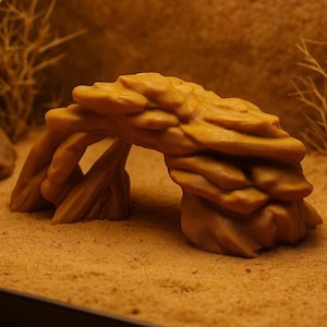 May include: A tan-coloured, artificial rock formation with an archway, designed for terrarium or aquarium use. The rock has a textured surface and is placed on a sandy substrate, with dried plants in the background.