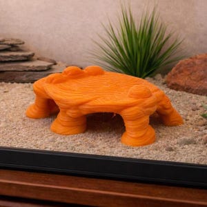 May include: An orange reptile hide with a textured surface and a bridge-like shape. The hide features four pillar-like legs and a textured top with two rounded protrusions. It sits on sand, with rocks and greenery in the background.